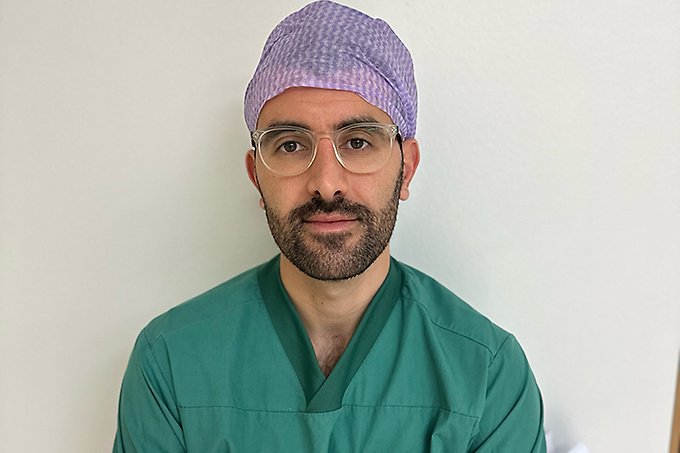 Halvkroppsbild på person i operationskläder, glasögon och mörkt skägg tittar in i kameran