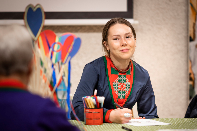 En person i samisk klädedräkt sitter vid ett bord med papper och penna. Närmast kameran skymtar ryggen på en annan  person i oskärpa.