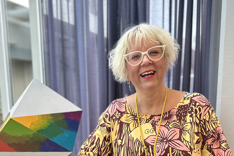 Närbild på leende person i kort blont hår, glasögon och mönstrad klänning