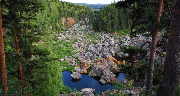 Bild på Döda fallet i Ragunda. I förgrunden syns omgivande tallar, i mitten ett mindre vattendrag och enorma stenrösen. I bakgrundsbilden syns mer skog och utsikt över en avlägsen sjö och fjälltoppar.