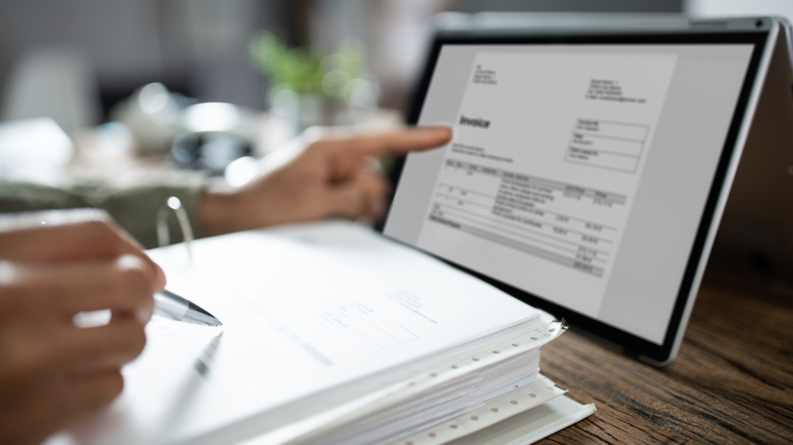 En hand pekar på en pekplatta med räkningar och ekonomiprogram på. Framför på bordet ligger en pärm med räkningar.