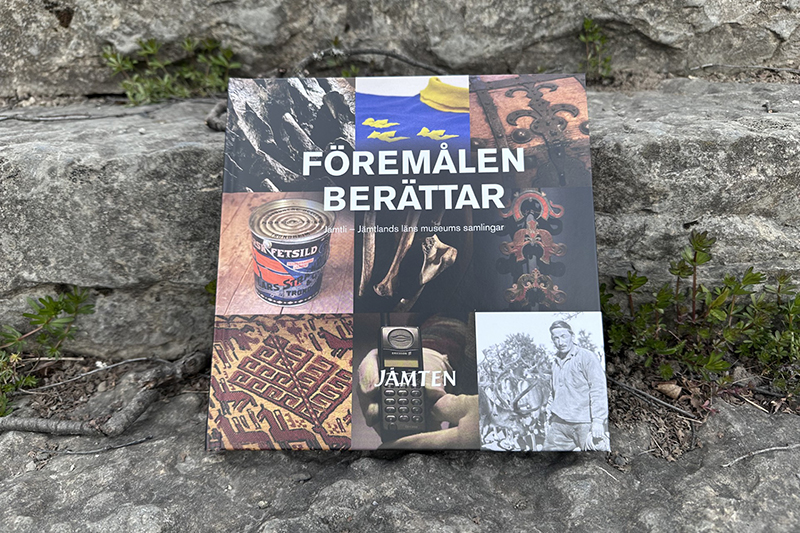En bok med färgglatt fotokollage på framsidan står lutad mot en stentrappa med gröna grästuvor runt sig