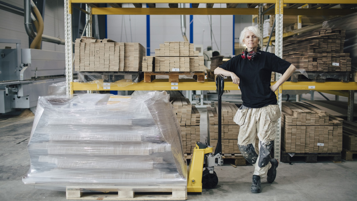 En företagare med arbetskläder står i ett lagervaruhus bredvid en gaffeltruck med inplastade produkter på pall.