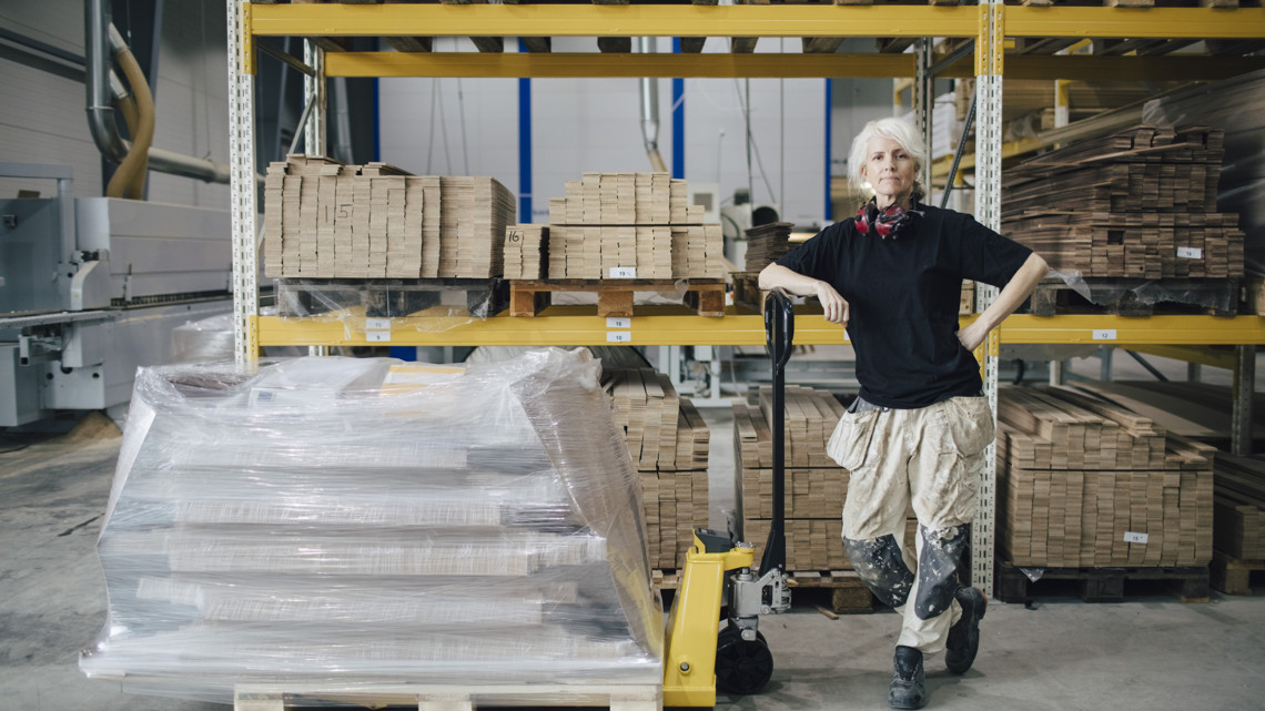 En företagare med arbetskläder står i ett lagervaruhus bredvid en gaffeltruck med inplastade produkter på pall.