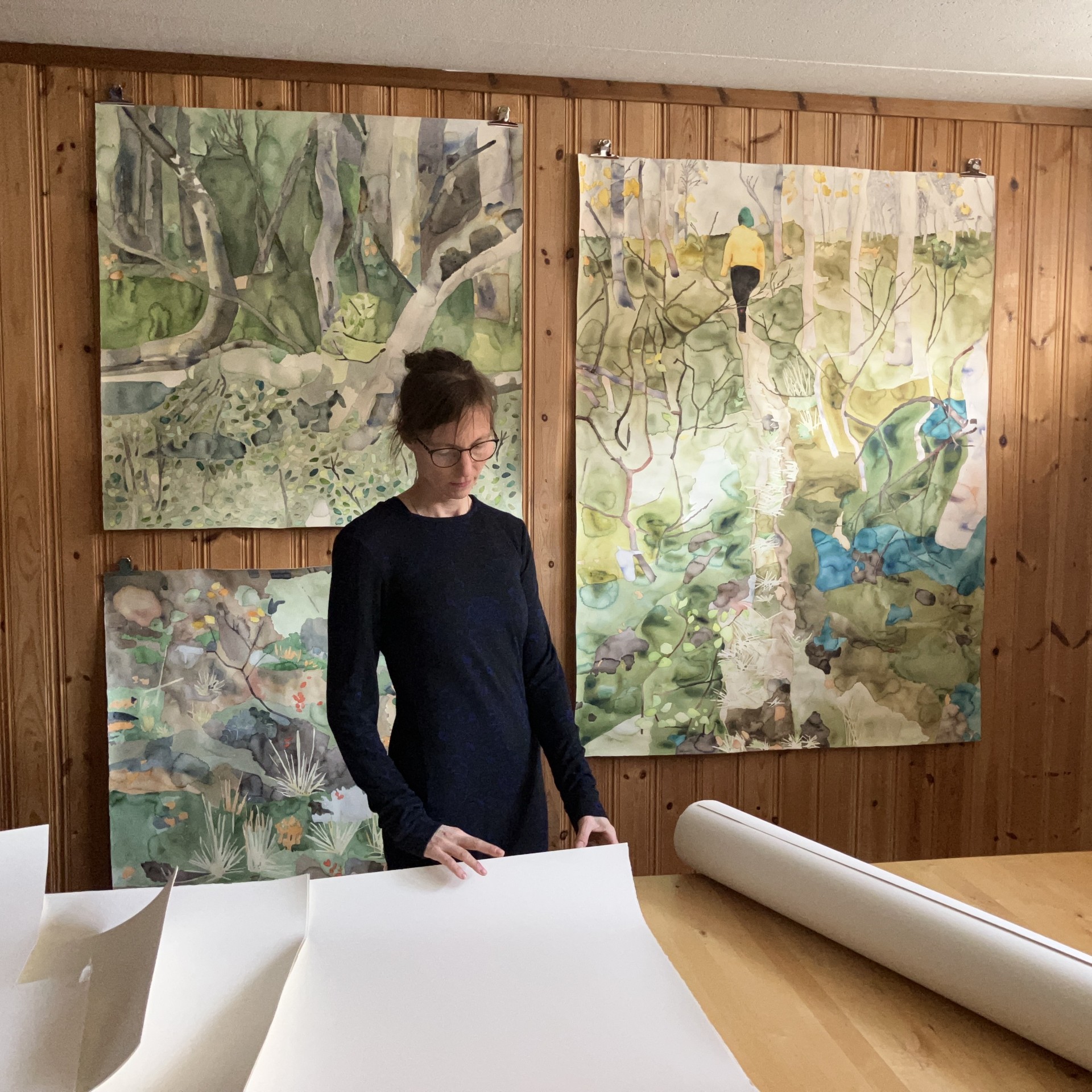 En person i svarta kläder står vid ett bord i en ateljé där färgglada målningar sitter på en trävägg.