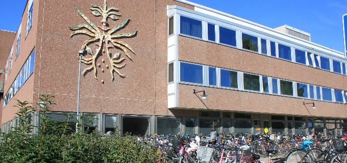 Miljöbild som visar en del av Östersunds sjukhus. På asfaltytan framför den rödbruna fasaden står en rad cyklar parkerade i cykelställ. Bredvid cykelställen finns gröna buskar.