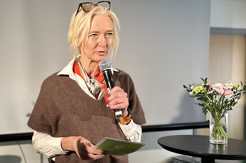 En person med blont hår och brun tröja med en mikrofon i sin hand