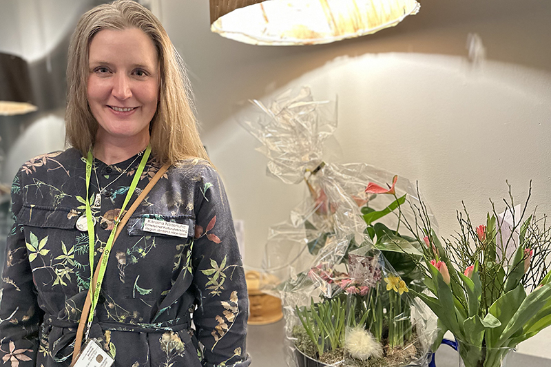 Person i blommig blus och långt hår ler mot kameran. Intill står flera vackra blomsterarrangemang