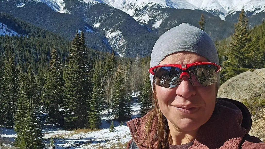 en person med solglasögon som står i skogen framför snöiga berg