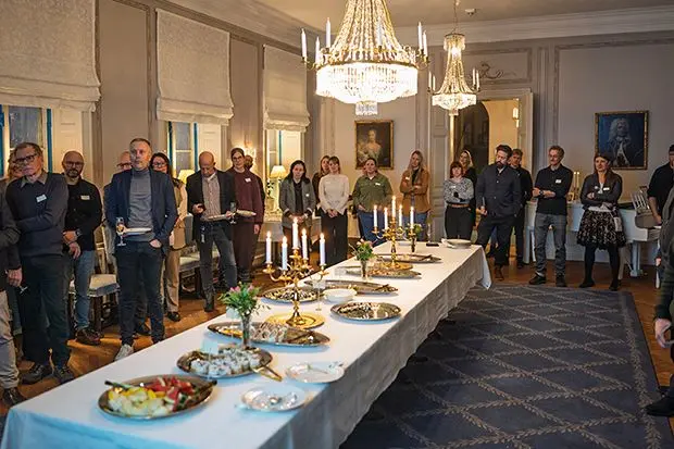 Ett antal personer står i ett rum. I mitten av rummet finns ett bord med uppläggningsfat. Personerna ser ut att lyssna på en talare som inte syns i bild. 