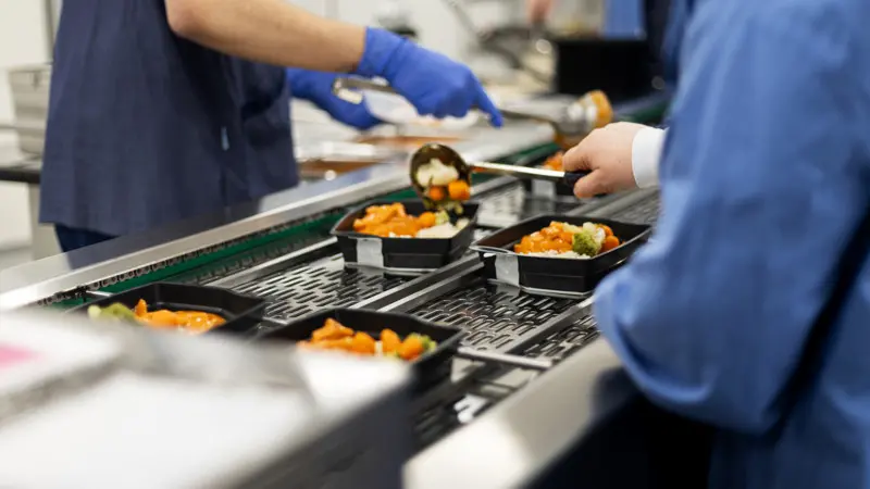 matlådor på ett rullband. Två personer lägger i mat i matlådorna.