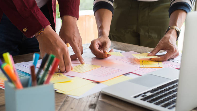 Närbild på två personers armar, när de samarbetar med post-its och skisser på ett skrivbord framför en bärbar dator.