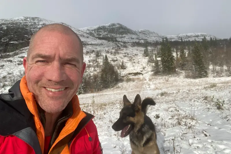 En person klädd i vinterjacka står på kalfjället på vintern i snön och ler in i kameran. I bakgrunden syns en schäfer och snöklädda fjäll.
