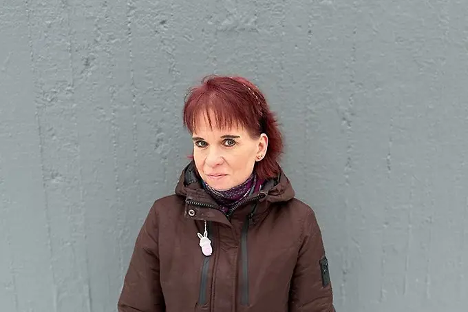 Person i vinterjacka och rött hår står mot en grå stenvägg och tittar in i kameran