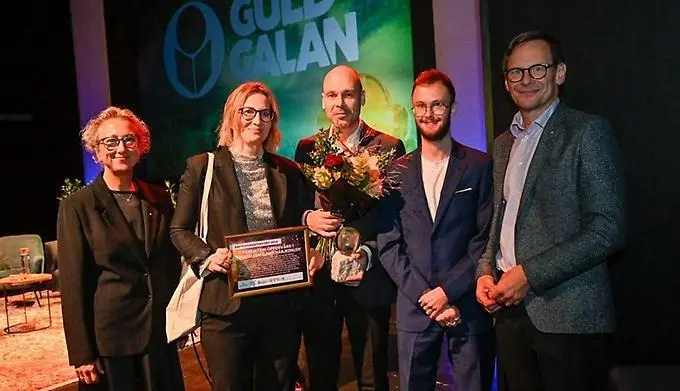 Fem personer står uppradade på en scen och tittar in i kameran. De två i mitten håller i ett diplom och blommor.