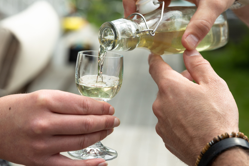 Närbild på en persons händer som håller i en liten glasflaska och häller upp dryck i ett snapsglas som en annan person håller i.