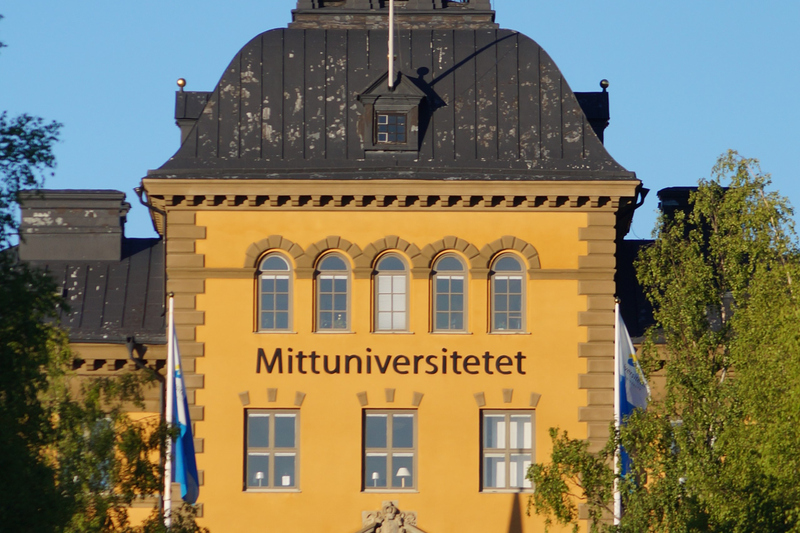 Framsidan av Mittuniversitetet med björkar  på vardera sida en klar sommardag.