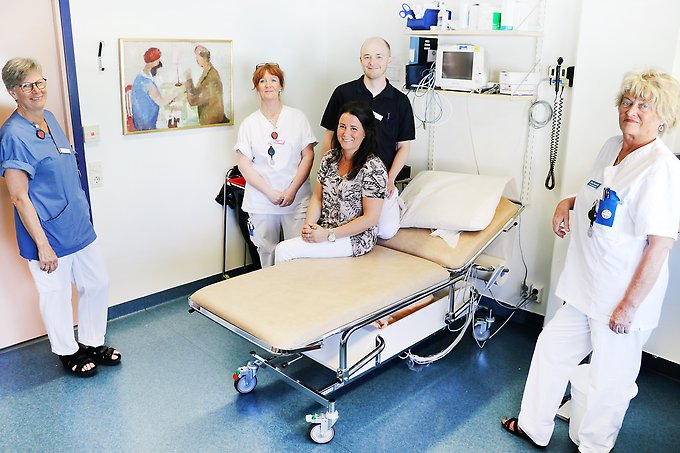 fem personer i ett vårdrum på en hälsocentral, en kvinna står och håller i dörren till rummet, en kvinna står med armen vilande på ett skrivbord, en kvinna och en man står bakom en brits och en kvinna sitter på britsen