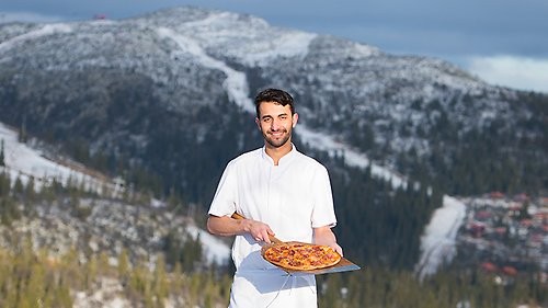 Porträttbild av en pizzabagare i vita bagarkläder som visar upp en pizza på sin stekspade, ståendes på fjället med ett halvsnöigt fjäll i bakgrunden.