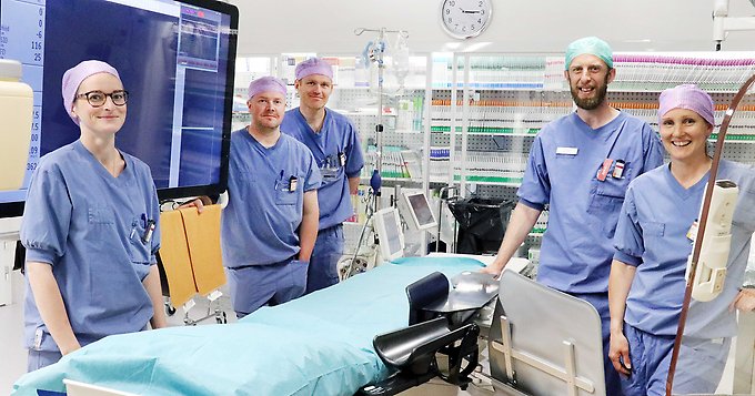 fem personer i blå vårdkläder och med hårnät står runt en brits i en operationssal