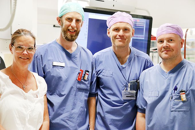 en civilklädd kvinnlig chef, två manliga läkare och en manlig sjuksköterska, klädda i blå vårdkläder och med hårnät, står bredvid varandra i en operationssal