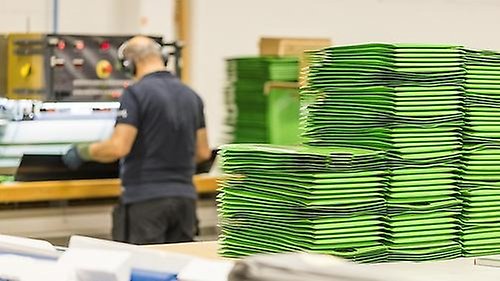 Fabriksarbetare står vid en maskin bredvid en hög med gröna kuvert som maskinen producerar.