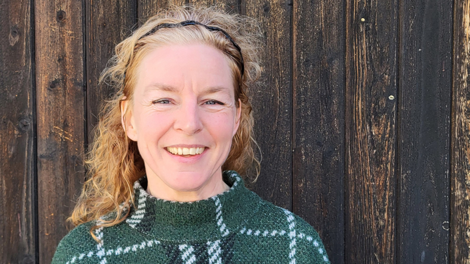 En leende person med långt lockigt hår och grön-vit tröja. I bakgrunden syns en mörk trävägg. 