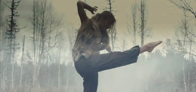 I mitten av fotografiet syns en dansare som står på ett ben. Dansaren har blicken mot marken och sträcker höger arm uppåt. I bakgrunden syns en gles glänta av björkar och granar. Fotografiet har ett filter i gula och bruna toner.